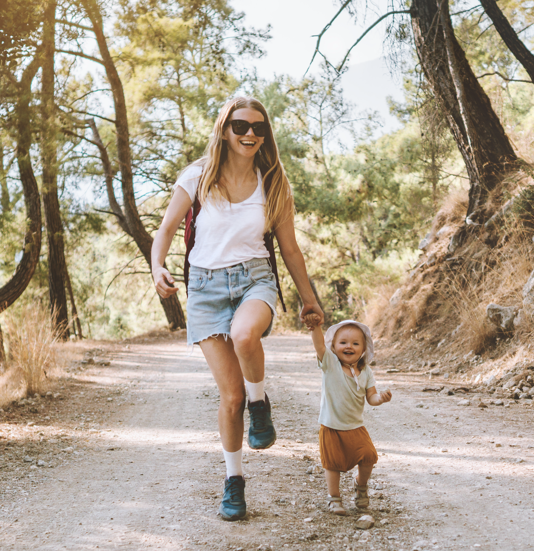 „Eine fröhliche Frau in Shorts und Sonnenbrille wandert auf einem Waldweg und hält die Hand eines lachenden Kleinkinds, das neben ihr läuft. Beide wirken glücklich und genießen einen sonnigen Tag in der Natur, umgeben von Bäumen und Hügeln.“