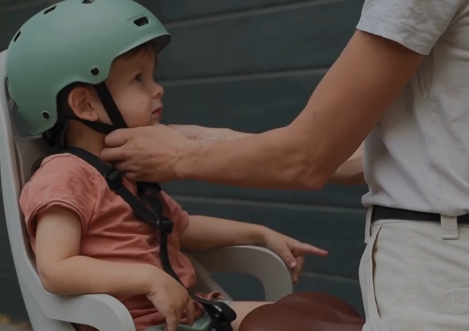„Ein fröhlicher brauner Hund mit grünem Geschirr läuft auf einem Waldweg auf die Kamera zu. Im Hintergrund wandern mehrere Menschen, darunter zwei Kinder und eine Frau, durch den sonnendurchfluteten Wald. Alle wirken entspannt und genießen die Natur.“