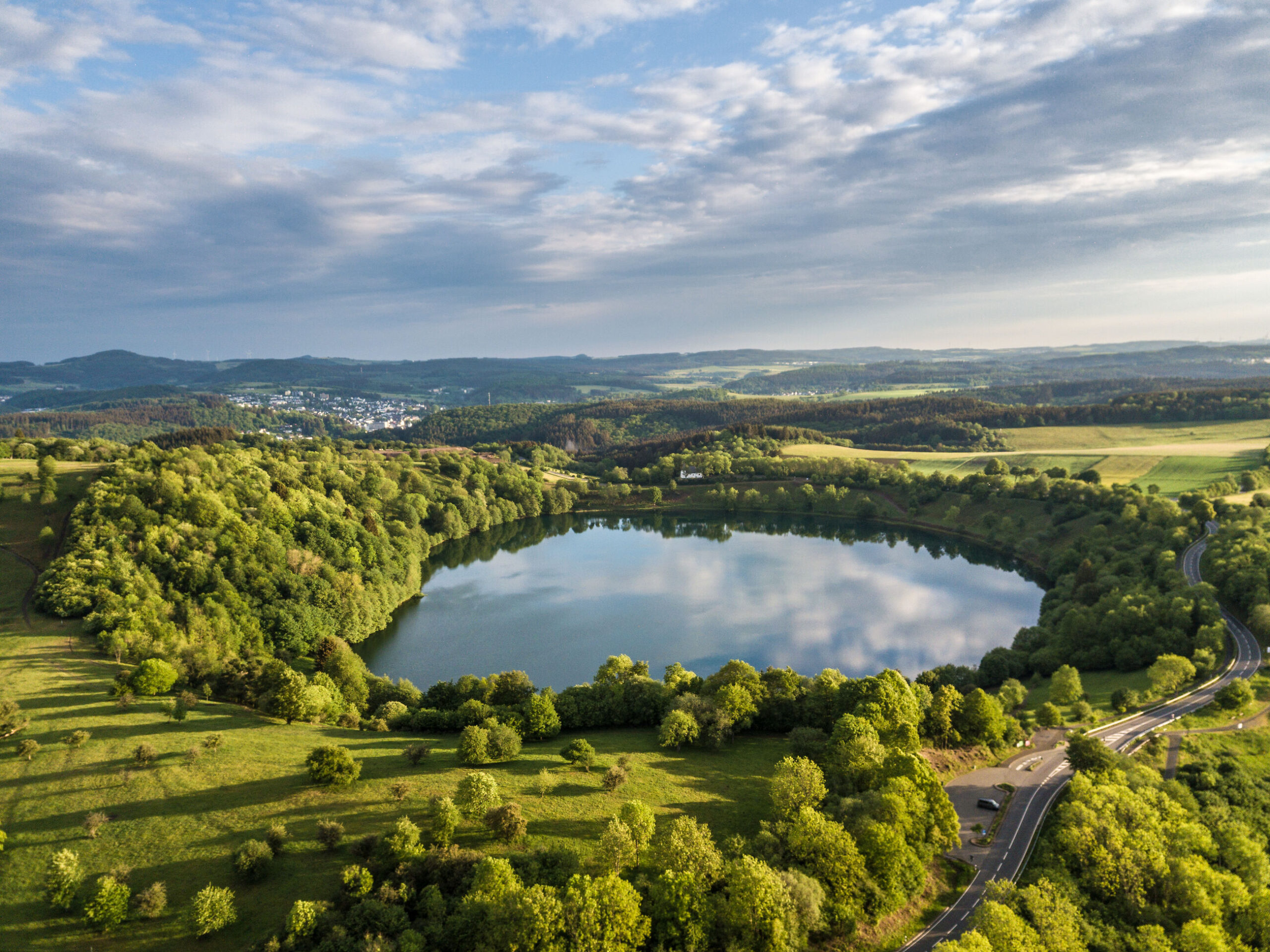 „Eine fröhliche Frau in Shorts und Sonnenbrille wandert auf einem Waldweg und hält die Hand eines lachenden Kleinkinds, das neben ihr läuft. Beide wirken glücklich und genießen einen sonnigen Tag in der Natur, umgeben von Bäumen und Hügeln.“